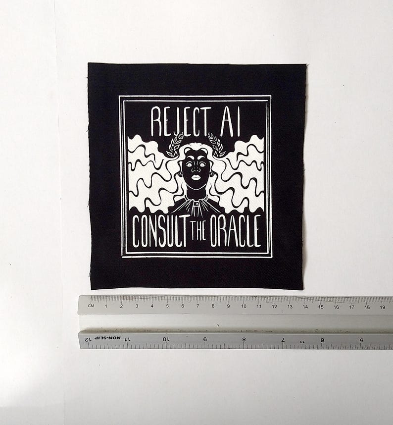 May include: A square black fabric patch featuring a white screen-printed graphic of a person with wavy hair wearing a laurel wreath. The text reads REJECT AI CONSULT THE ORACLE. A ruler placed below shows the patch is approximately 14 cm wide.