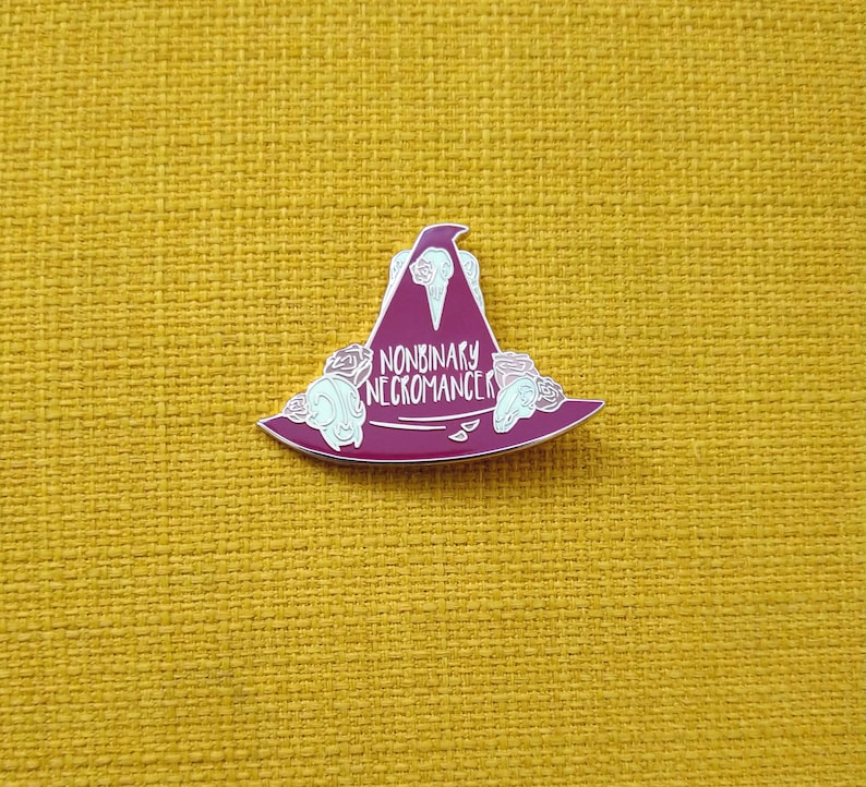 May include: A red enamel pin with a witch's hat and the text "Nonbinary Necromancer" in white.