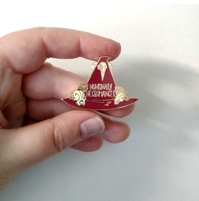 May include: A red enamel pin shaped like a witch's hat with gold accents. The hat has a skull and crossbones design and the text "Nonbinary Necromancer" on the front.