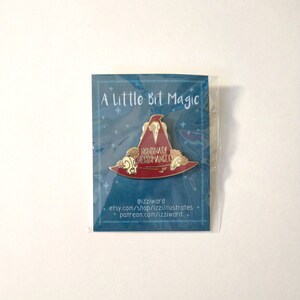 May include: A red and gold enamel pin featuring a witch's hat with the text "Nonbinary Witchromancer" on it. The pin is packaged on a blue card with the text "A Little Bit Magic" and the artist's social media handles.