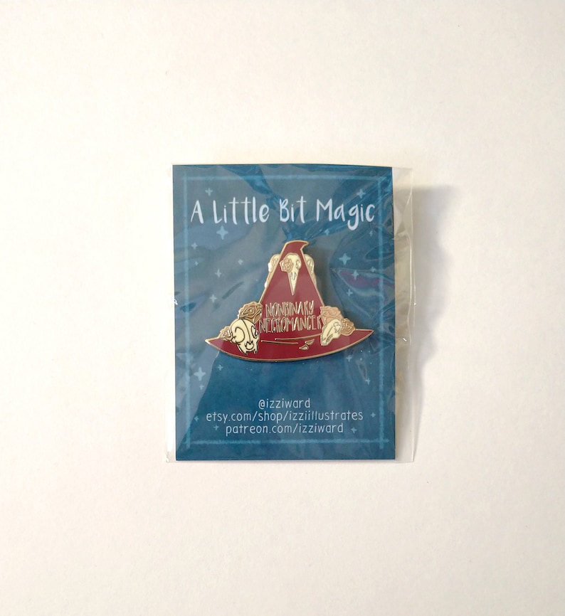 May include: A red and gold enamel pin featuring a witch's hat with the text "Nonbinary Witchromancer" on it. The pin is packaged on a blue card with the text "A Little Bit Magic" and the artist's social media handles.