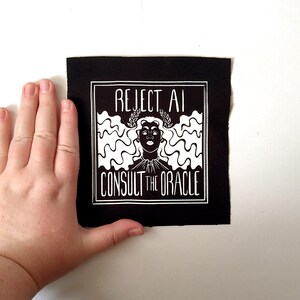 May include: A square black fabric patch featuring a white screen-printed graphic of a person with long wavy hair wearing a laurel wreath. The text REJECT AI is printed above the head and CONSULT THE ORACLE is printed below the graphic design.