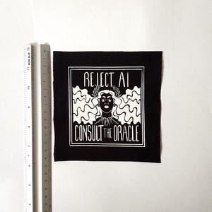 May include: A square black fabric patch featuring a white screen-printed graphic of a person with wavy hair and a laurel wreath. The text reads REJECT AI CONSULT THE ORACLE. A metal ruler is placed alongside, showing the patch is 12 cm in height.