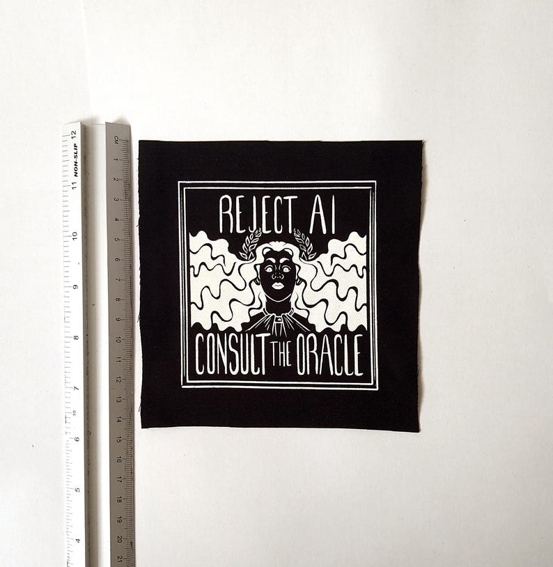 May include: A square black fabric patch featuring a white screen-printed graphic of a person with wavy hair and a laurel wreath. The text reads REJECT AI CONSULT THE ORACLE. A metal ruler is placed alongside, showing the patch is 12 cm in height.