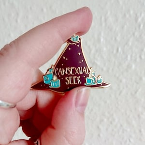 May include: Gold-toned enamel pin featuring a purple witch's hat with teal candles and the text "Pansexual Seek" in gold.