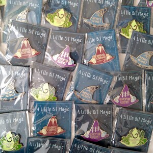 May include: A collection of enamel pins featuring various witch hat designs with different slogans. The slogans include "Nonbinary Necromancer", "Genperfluid Alchemist", and "Agent of the Arcanist". The pins are packaged in clear plastic bags with a black background and the words "A Little Bit Magic" printed on the front.