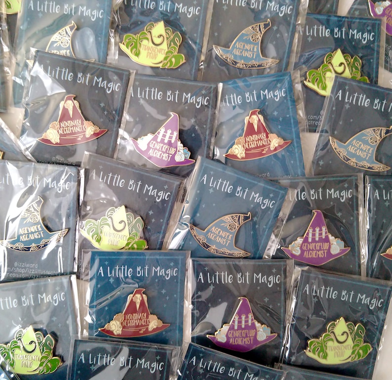 May include: A collection of enamel pins featuring various witch hat designs with different slogans. The slogans include "Nonbinary Necromancer", "Genperfluid Alchemist", and "Agent of the Arcanist". The pins are packaged in clear plastic bags with a black background and the words "A Little Bit Magic" printed on the front.