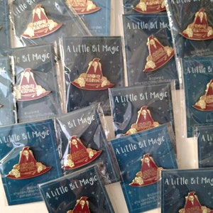 May include: A collection of enamel pins featuring a red and gold wizard hat with the text "Nominally Acceptable Wizard" on it. The pins are packaged in clear plastic bags with a blue background card that reads "A Little Bit Magic".