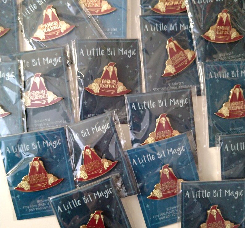 May include: A collection of enamel pins featuring a red and gold wizard hat with the text "Nominally Acceptable Wizard" on it. The pins are packaged in clear plastic bags with a blue background card that reads "A Little Bit Magic".