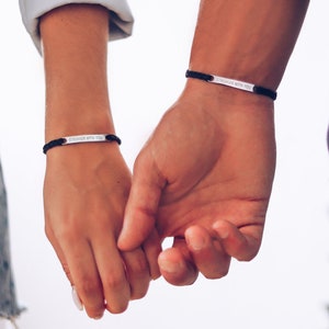May include: Two people holding hands, each wearing a black bracelet with a silver plate that reads "Stronger with you".