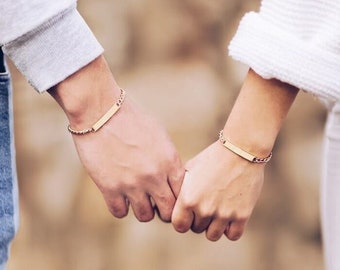 以下が含まれることがあります： 2 人が手を繋いでおり、それぞれが小さな長方形のタグが付いた金のチェーンブレスレットをしています。