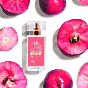 May include: A clear perfume bottle with a pink label featuring the words "Sugar Sweet" and "Bom Dia Bright" in gold lettering. The bottle is surrounded by fresh, halved plums on a white background. The perfume bottle contains 30ml (1 fl. oz.) of organic perfume.