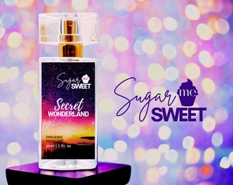 May include: A clear perfume bottle with a gold-colored sprayer and a clear cap. The label reads "Sugar me SWEET Secret Wonderland Organic Perfume 30ml | 1 Fl. oz." The bottle sits on a black stand against a bokeh background.