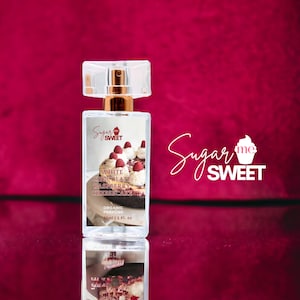 May include: A rectangular perfume bottle with a clear, faceted cap and gold-colored neck. The label features the text "Sugar me Sweet" and "White Chocolate Raspberry Cheesecake." The bottle contains 1 fl oz of organic perfume, set against a deep red background.