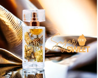 May include: A clear perfume bottle with a gold-colored atomizer and a clear cap. The label reads "Bak Lava" with images of baklava and honey. The words "Sugar Me Sweet" are in a script font on the right side of the bottle. The bottle is reflected on a shiny surface.