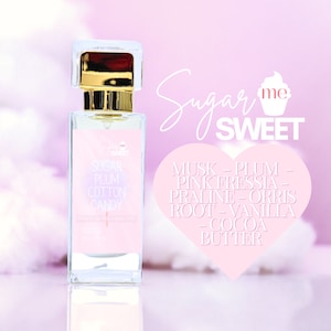 May include: A rectangular perfume bottle with a clear glass body and a gold-colored cap. The label reads "Sugar Plum Cotton Candy" with a list of fragrance notes. The background is a soft pink with the words "Sugar me SWEET".