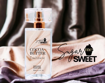 May include: A rectangular perfume bottle with a clear cap and gold-colored accents. The label reads "COCOA BUTTER Cashmere" and "ORGANIC PERFUME 30ml (1 Fl. oz)". The background features draped fabric in shades of brown and purple.