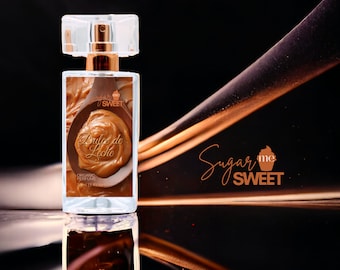 May include: A clear glass perfume bottle with a gold-colored top and a rectangular label featuring a wooden spoon filled with caramel. The label reads "Dulce de Leche" and "Organic Perfume". The brand name "Sugar me Sweet" is also visible.