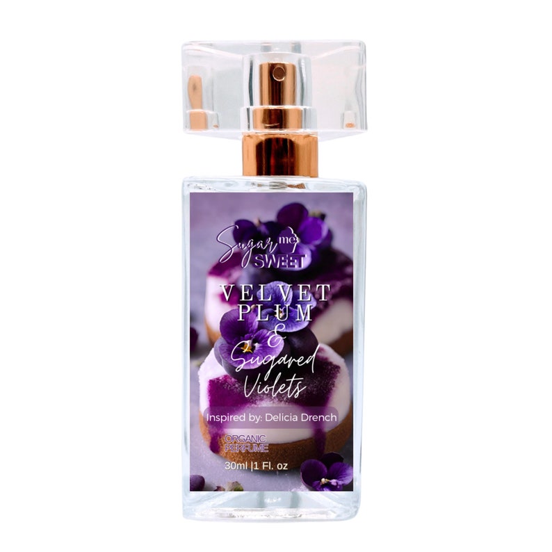 May include: A clear perfume bottle with a gold-colored atomizer and a rectangular label. The label features the text "Sugar Me Sweet" and "Velvet Plum & Sugared Violets" over a blurred image of a dessert with purple flowers. The bottle contains 1 fl oz.