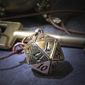 May include: A metallic twenty-sided die pendant with black engraved numbers, suspended from a thin brown cord. The die features a polished finish and is positioned in front of a large, antique-style brass key with a rectangular bow and bit.