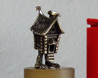May include: A small, detailed bronze figurine of a house with legs, standing on a brown pedestal. The figurine is next to a red lighter for scale.