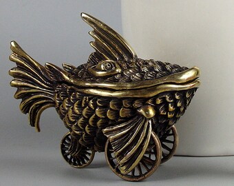 May include: A gold-coloured metal sculpture of a fish with wheels. The fish has a detailed design and appears to be a decorative object. The sculpture is sitting on a white surface next to a lighter with the words "Peace, Love, Weed" on it.