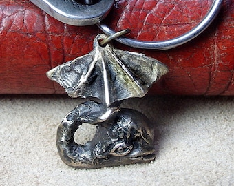 May include: A bronze-coloured keyring with a sea creature pendant and wing-like structure. The pendant hangs from a silver key ring, with keys in the background. The background is a textured, reddish-brown surface.