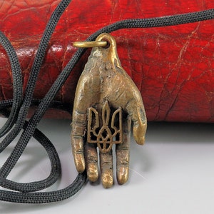 Pode incluir: Um pingente de bronze em forma de mão segurando o símbolo do tridente ucraniano. O pingente está em um cordão preto e está sobre uma superfície de couro vermelho.