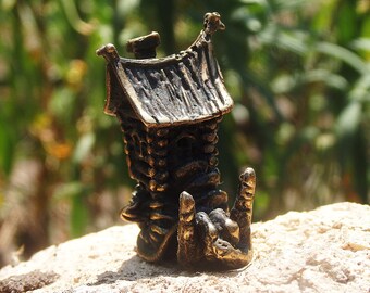 May include: A small, bronze figurine of a house with a chimney and a pointed roof. The house is perched on a rock with a whimsical, almost cartoonish, design.