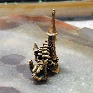 Puede incluir: Una pequeña escultura de metal de color bronce detallada de un castillo con una aguja puntiaguda. El castillo está intrincadamente diseñado con una escalera de caracol y múltiples torres.