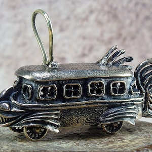 May include: A silver-coloured pendant shaped like a bus with wings. The bus has a detailed front with a toothy grin and windows. The wings are made of intricate metalwork.