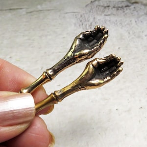 May include: Two gold-toned metal bottle stoppers shaped like open hands with the palms facing up.
