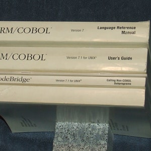 Könnte beinhalten: Drei weiße Bücher, die übereinander gestapelt sind. Das oberste Buch trägt den Titel "RM/COBOL Sprachreferenzhandbuch" und die Versionsnummer "7". Das mittlere Buch trägt den Titel "RM/COBOL Benutzerhandbuch" und die Versionsnummer "7.1 für UNIX". Das unterste Buch trägt den Titel "CodeBridge Aufruf von Nicht-COBOL-Unterprogrammen" und die Versionsnummer "7.1 für UNIX".