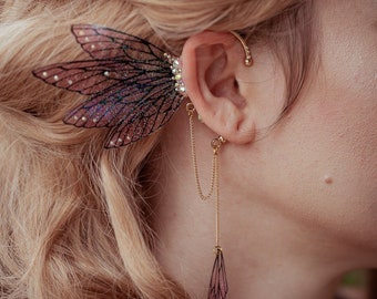 Puede incluir: Un detallado pendiente de oreja con un diseño delicado en forma de ala con tonos rosas y morados. El pendiente está adornado con cristales brillantes y una cadena colgante con un dije de ala a juego. La joya es dorada.