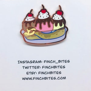 May include: A gold-tone enamel pin depicting a banana split with three scoops of ice cream, chocolate sauce, whipped cream, cherries, and a snake peeking out from under the banana.