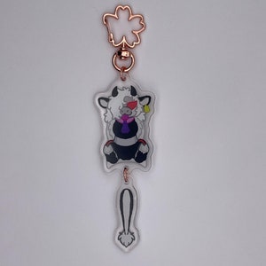 May include: A white and black furry keychain with a purple bow tie and a gold bell. The keychain is attached to a gold metal flower-shaped charm.