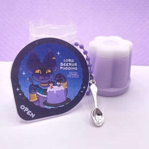 Può includere: Un contenitore di budino viola e bianco con un coperchio a forma di fiore. Un portachiavi con un'immagine di cartone animato di un gatto viola con una corona e due figure più piccole, un cucchiaio e il testo "Lord Beerus Pudding Taro Flavor" e "Open".