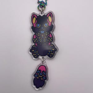 May include: An acrylic keychain featuring a black cat character with pink inner ears, blue eyes, and a patterned chest and paws with yellow, pink, and blue geometric shapes. A separate smaller tail charm hangs below, attached by a metal ring to a star-shaped clasp.