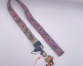Op de afbeelding: Een kleurrijk koord met een roze, groen en zwart patroon. Het koord heeft een zwarte clip en een roze en zwarte hanger eraan.