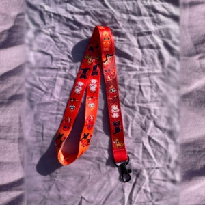 May include: Red lanyard with a repeating pattern of cartoon foxes, some with orange fur and some with white fur. The lanyard has a black metal clasp.