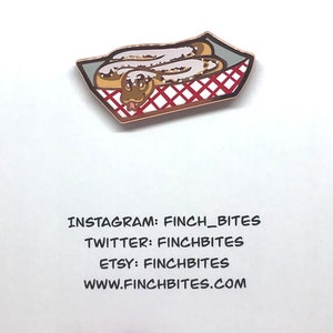 May include: A gold-coloured enamel pin featuring a snake in a red and white checked takeaway container. The pin has the text "INSTAGRAM: FINCH_BITES TWITTER: FINCHBITES ETSY: FINCHBITES WWW.FINCHBITES.COM".