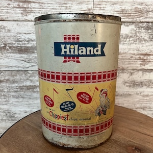 May include: Vintage Hiland potato chip tin. The cylindrical tin features a white background with red and blue accents. The Hiland logo is prominently displayed, along with text and illustrations promoting different chip flavors. The tin is slightly worn, adding to its vintage appeal.