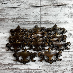 May include: A collection of antique bronze drawer pulls with ornate designs. The pulls have a dark, aged finish and feature intricate scrollwork and decorative details. They are arranged on a weathered, light-colored wooden surface.