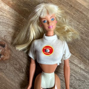 May include: A vintage doll with blonde hair and blue eyes, wearing a white crop top with a "Baywatch Lifeguard" logo. The doll has pink earrings and is posed against a wooden surface.