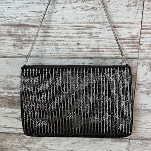 May include: Black rectangular beaded clutch with a silver chain strap. The bag is covered in vertical rows of black and silver beads. The zipper is black, and the chain is silver. The background is a light-colored, weathered wood.