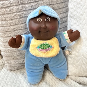 May include: A plush doll with dark skin, brown eyes, and rosy cheeks, wearing a light blue hooded onesie and a yellow bib. The bib features embroidered text that reads Cabbage Patch BABYLAND in green and pink lettering with a cabbage graphic.