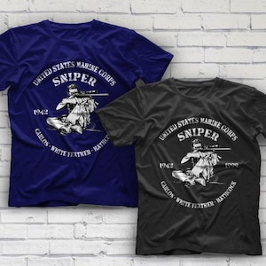 May include: Two t-shirts with a white graphic print of a United States Marine Corps sniper. The text on the graphic reads "United States Marine Corps Sniper 1942-1999 Carlos White Feather Hathcock". The t-shirt on the left is navy blue and the t-shirt on the right is black.