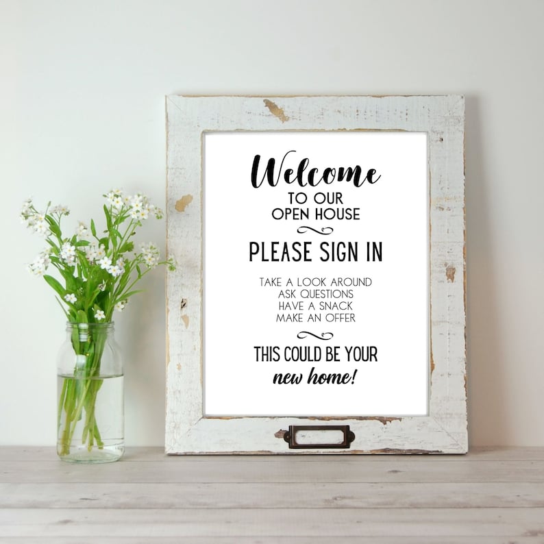 Op de afbeelding: Een wit bord in een rustiek houten frame met de tekst "Welkom bij onze open huis. Meld u aan. Neem een kijkje, stel vragen, neem een hapje, doe een bod. Dit zou uw nieuwe huis kunnen zijn!"