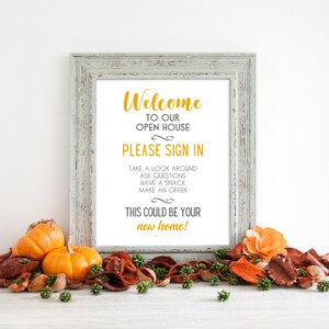 Op de afbeelding: Een wit ingelijst bord met de tekst "Welkom bij onze open huis. Meld u aan. Neem een kijkje, stel vragen, neem een hapje, doe een bod. Dit zou uw nieuwe huis kunnen zijn!" in zwarte en gouden tekst. Het bord staat op een tafel met herfstdecoraties.