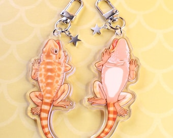 May include: Two bearded dragon keychains with silver keyrings and star charms. One keychain features a light orange bearded dragon with orange and white stripes on its tail. The other keychain features a light pink bearded dragon with orange and white stripes on its tail.
